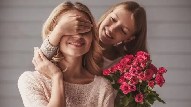 Pretty teenage daughter is holding flowers and covering her mom's eyes while making a surprise, both are smiling per sabattini