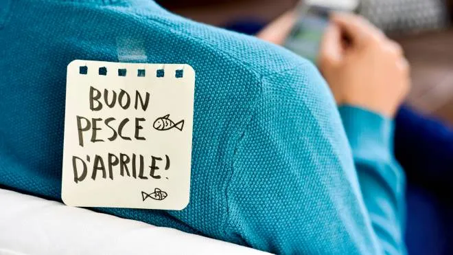 closeup of a young caucasian man seen from behind using his smartphone, with a note with the text buon pesce d aprile, happy april fools day in italian, attached with tape to his back