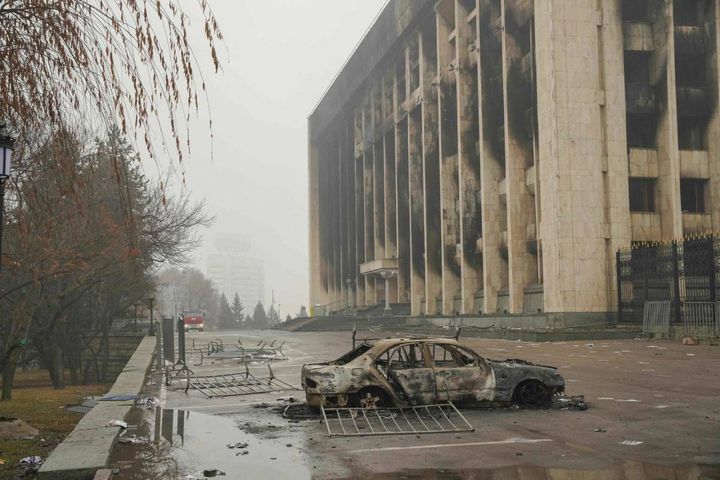 Un'auto distrutta dal fuoco di fronte a un palazzo amministrativo incendiato